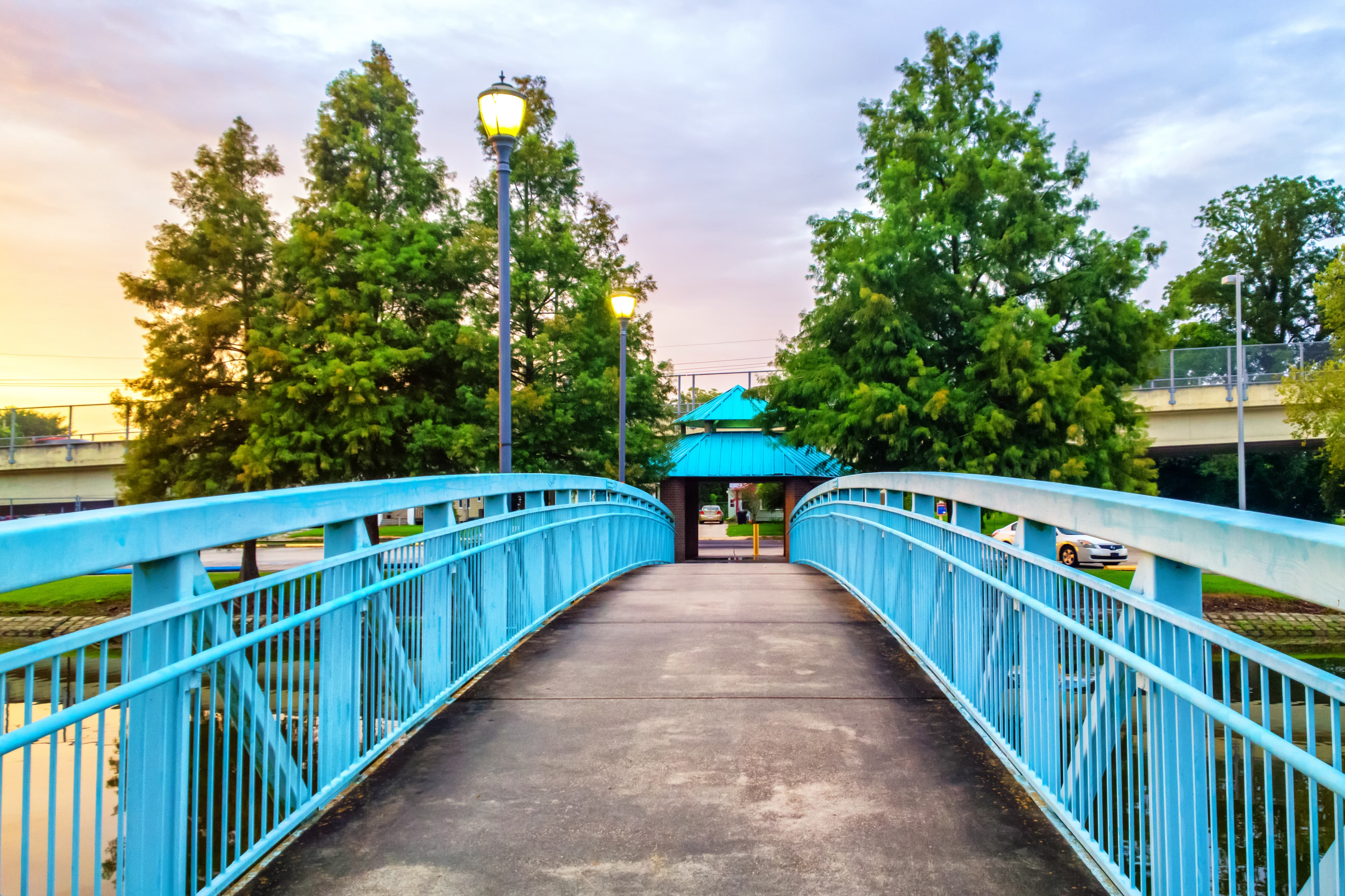 A picturesque blue pedestrian bridge leading into a park, framed by lush trees and warm streetlights, creating a peaceful and inviting scene at dusk.