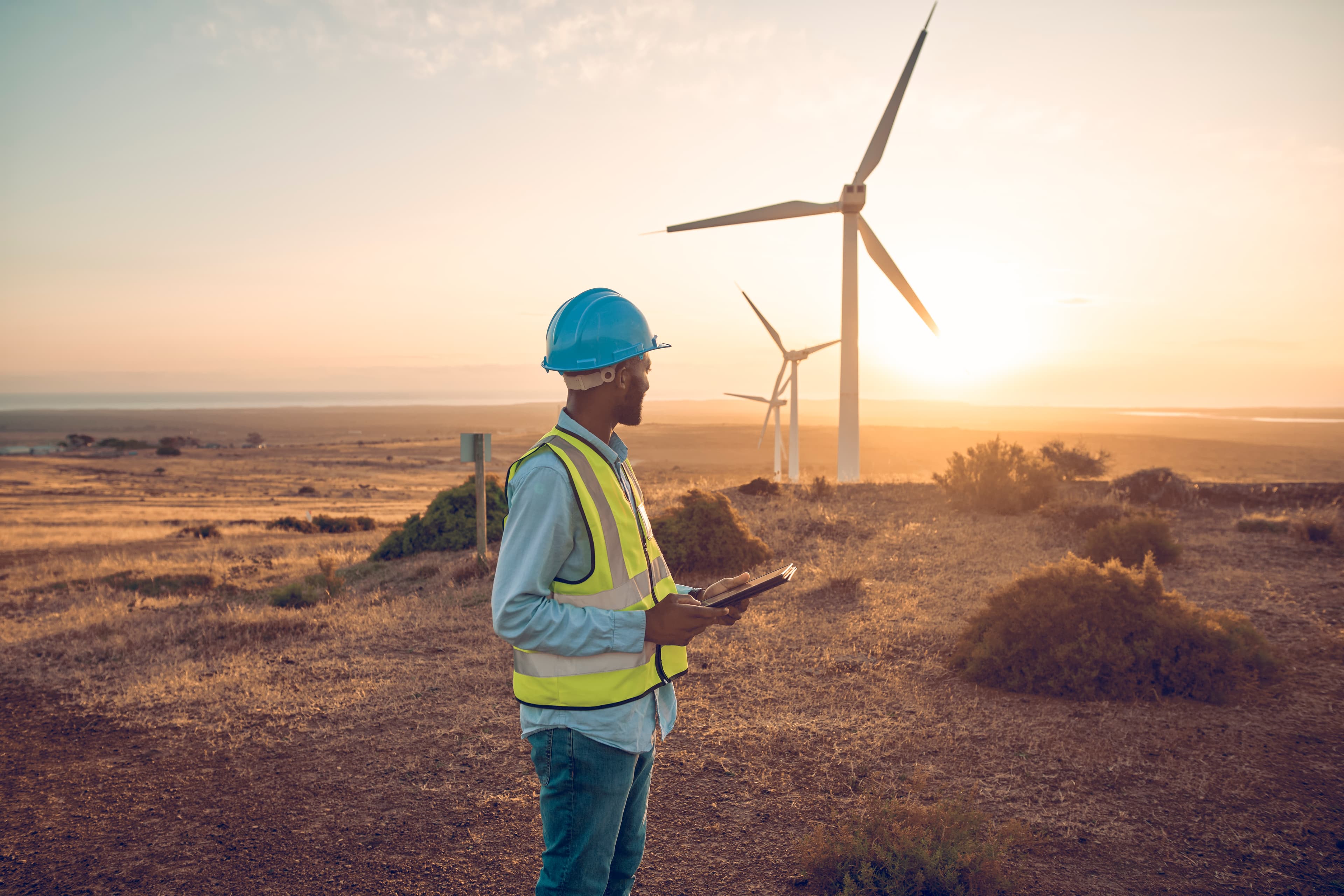 Engineer in safety gear conducting data collection at an onshore wind farm during sunset, with wind turbines in the background.