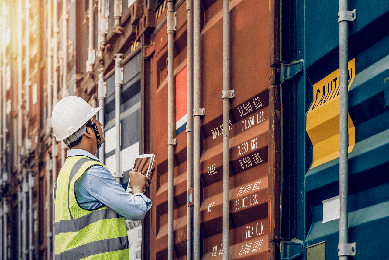 A person in a hard hat and high-visibility vest inspects shipping containers with a tablet, likely for logistics or safety checks at a port facility.