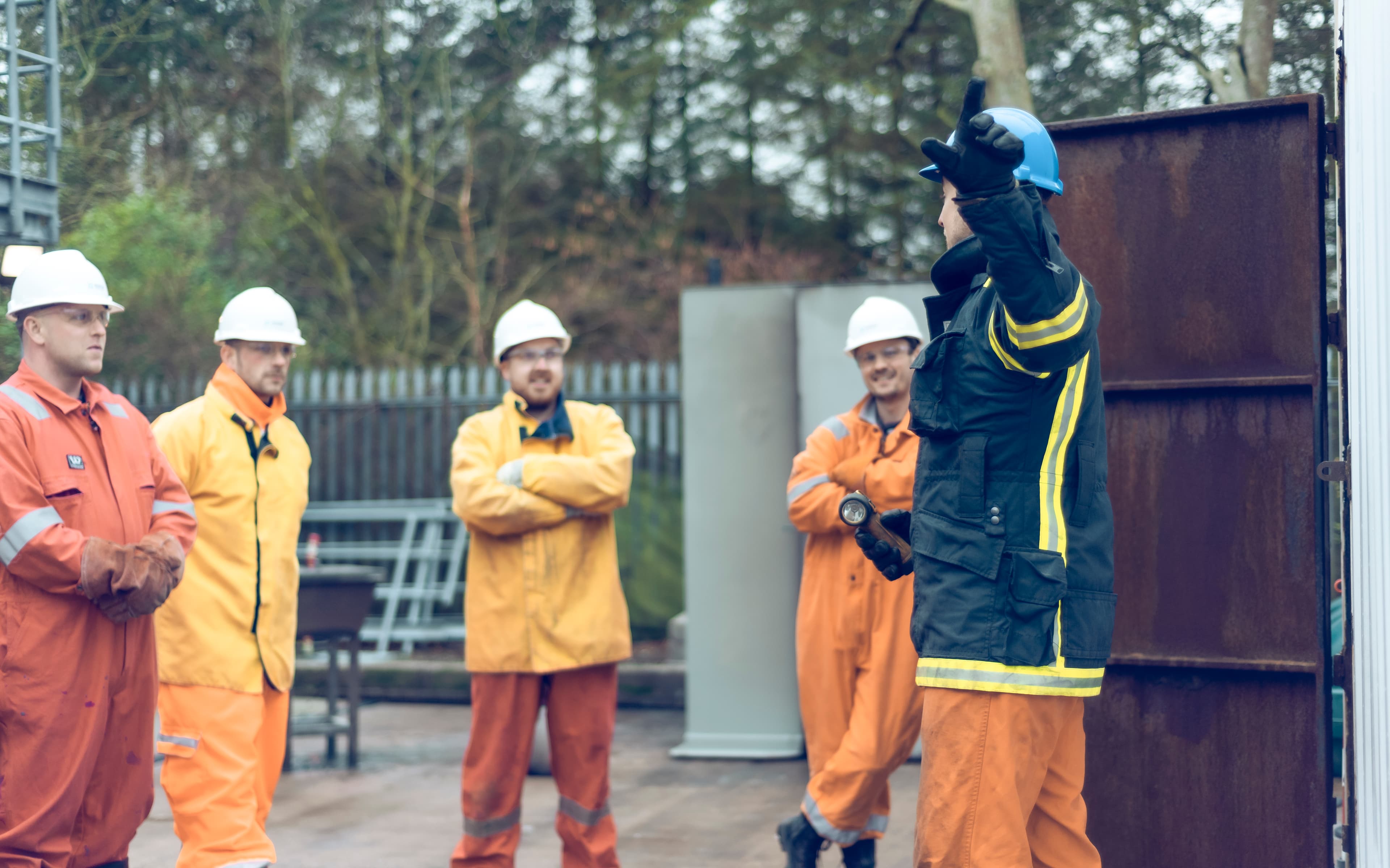 group of workers wearing safety gear, including hard hats and fire-resistant suits, gathered outdoors for a training session.
