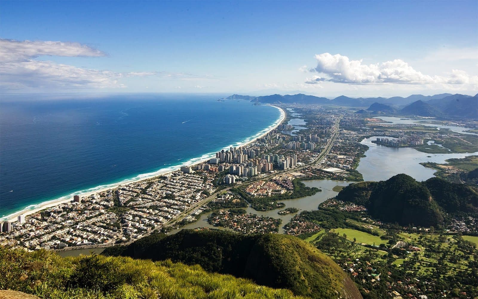 A panoramic view of Barra da Tijuca in Rio de Janeiro, showcasing its expansive coastline, urban landscape, and lush mountainous surroundings, blending natural beauty with modern development.