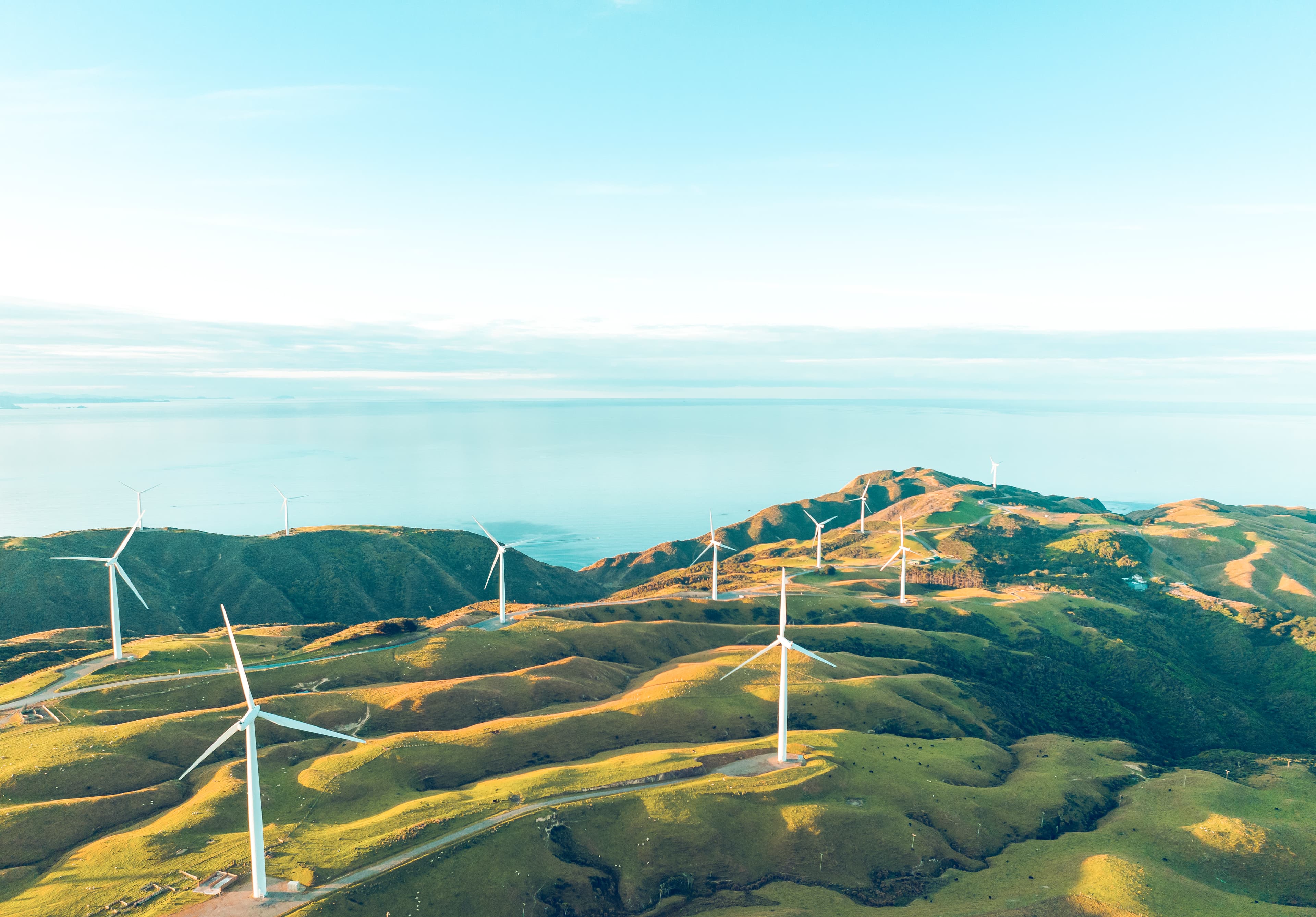 Aerial view of an onshore wind farm with multiple wind turbines spread across rolling green hills near the coastline.